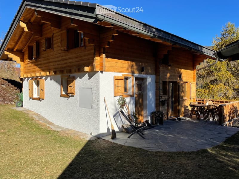 Two-story wooden house with white walls, wooden shutters, and a stone patio with outdoor furniture.