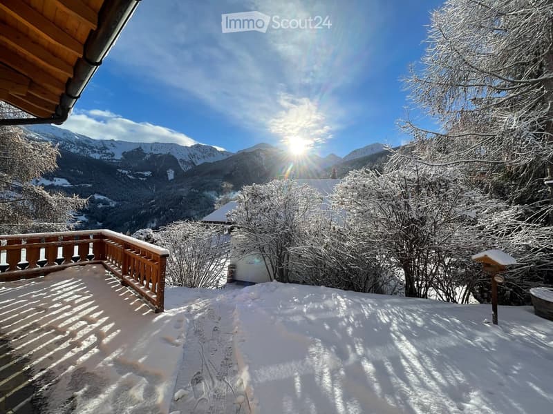Snow-covered balcony with wooden railing, overlooking a snowy mountain landscape and trees.