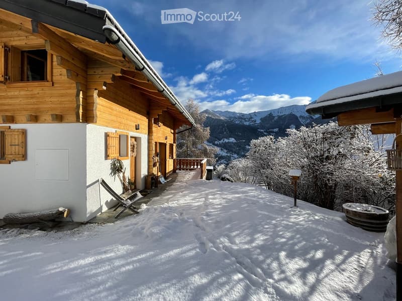 A wooden chalet with a snow-covered roof and a pathway leading to a balcony overlooking a snowy mountain landscape.