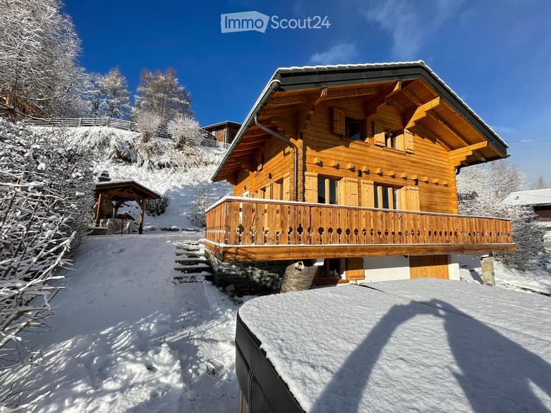 A large wooden chalet with a balcony, situated in a snowy mountain landscape with surrounding trees.