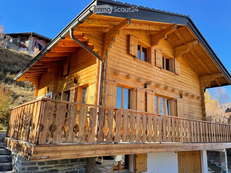A two-story wooden house with a balcony, large windows, and wooden shutters. The house features a raised ground floor and a stone base.