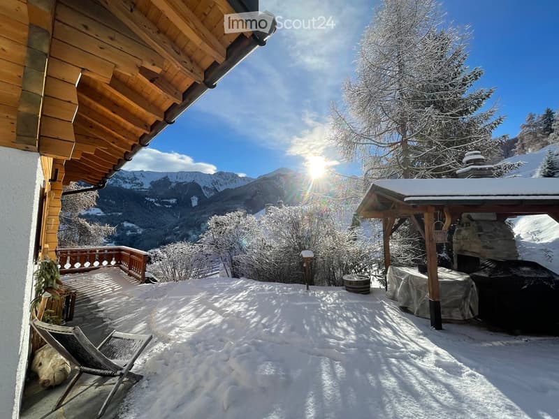 Snow-covered outdoor area with wooden deck, lounge chair, and scenic mountain view.