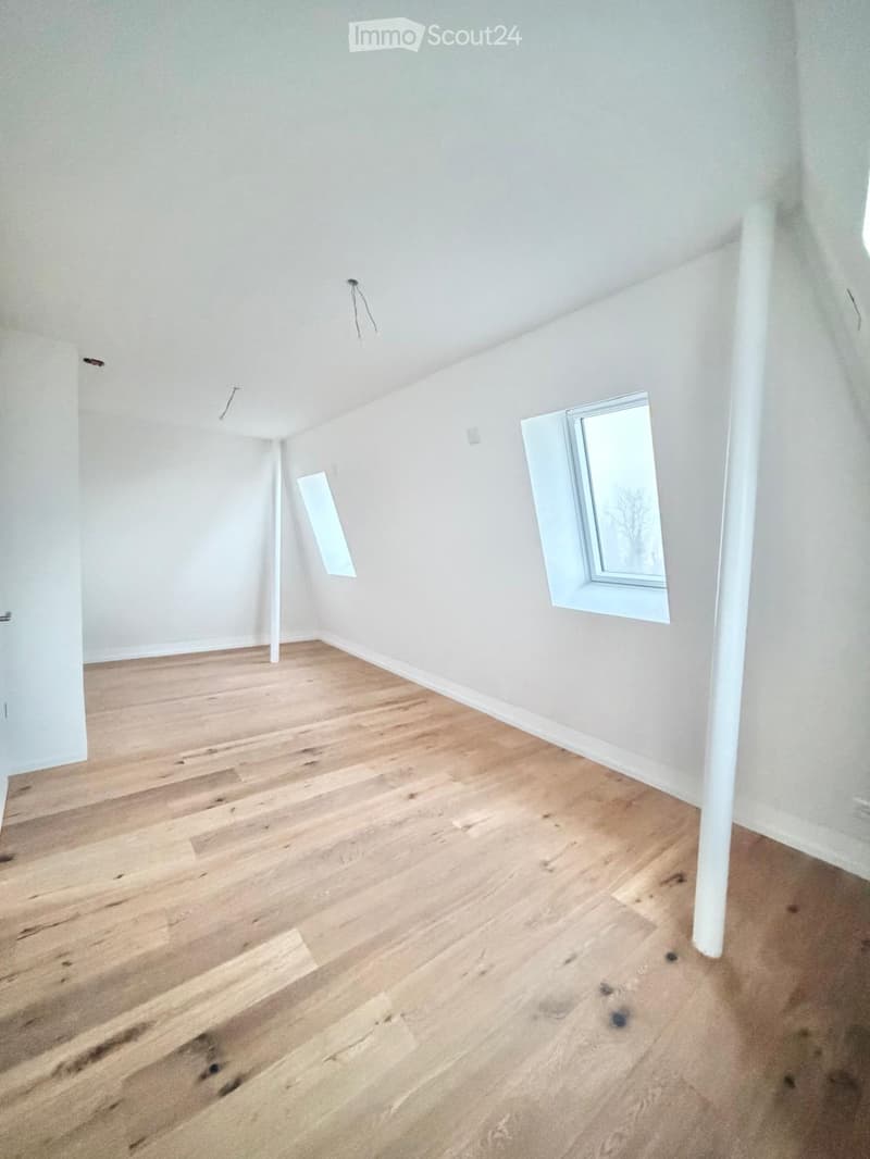 Empty room with light wooden flooring, white walls, and two windows with white frames. The room has a slanted ceiling typical of an attic space.