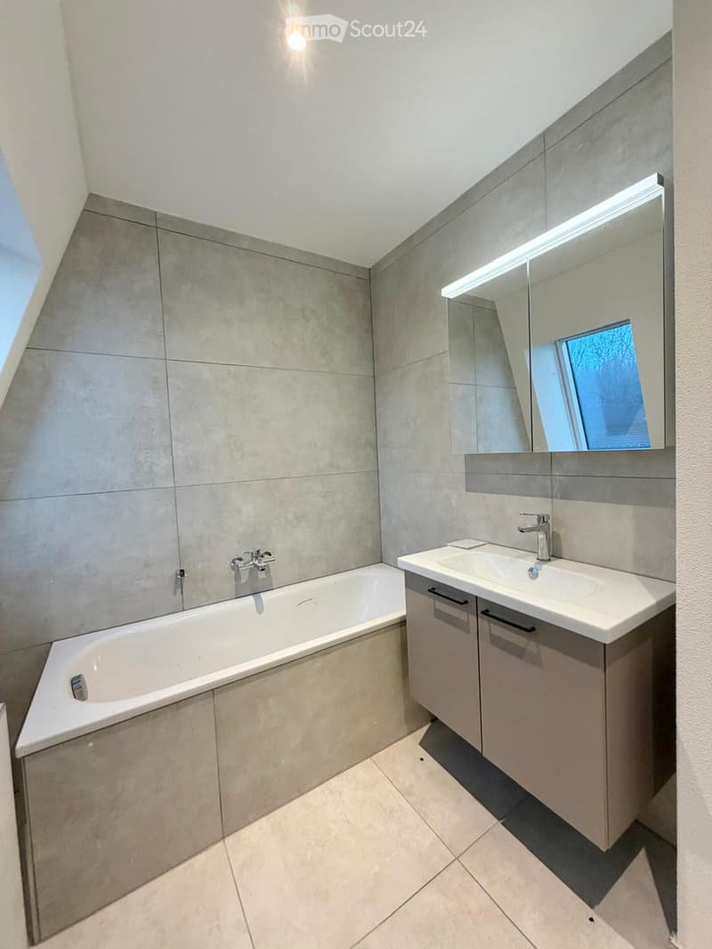 Modern bathroom with a bathtub, sink, and large mirror with built-in lighting. Grey tiles and sleek cabinetry.