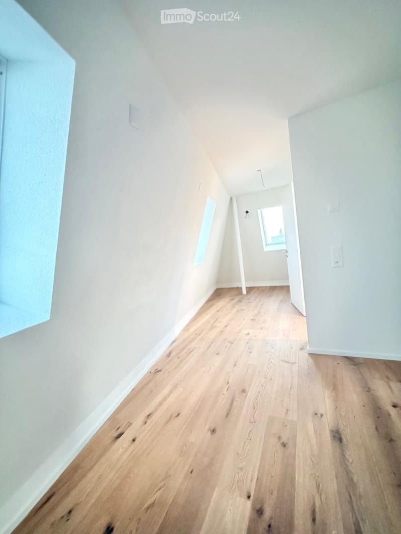Empty hallway with light wooden flooring and white walls, featuring a small window at the end and a door leading to another room.