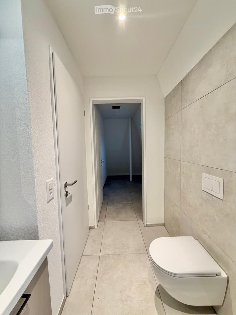 Modern bathroom with white tiles, a toilet, a sink, and a hallway leading to another room.