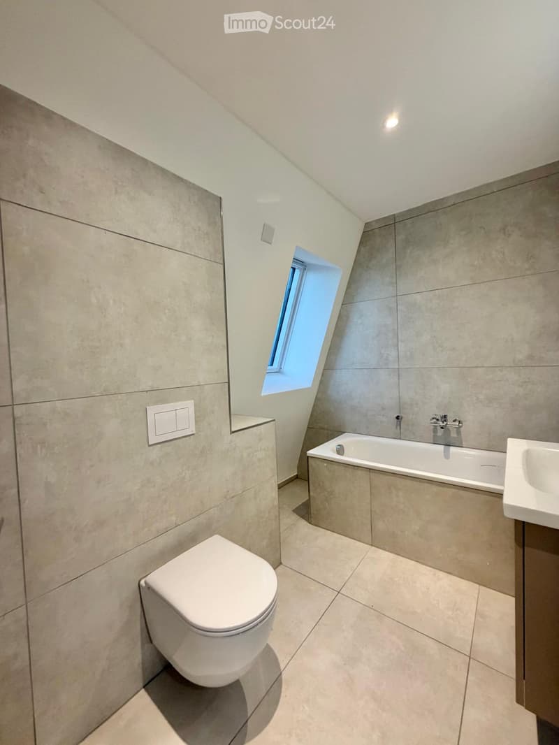 Modern bathroom with beige tiled walls and floor, featuring a toilet, bathtub, sink, and a skylight window providing natural light.