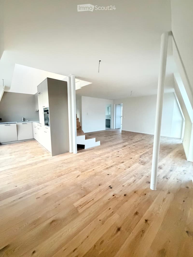Empty room with light wooden flooring, white walls, and a staircase leading to another level. The room has a modern, minimalist design with visible kitchen cabinets and appliances in the background.