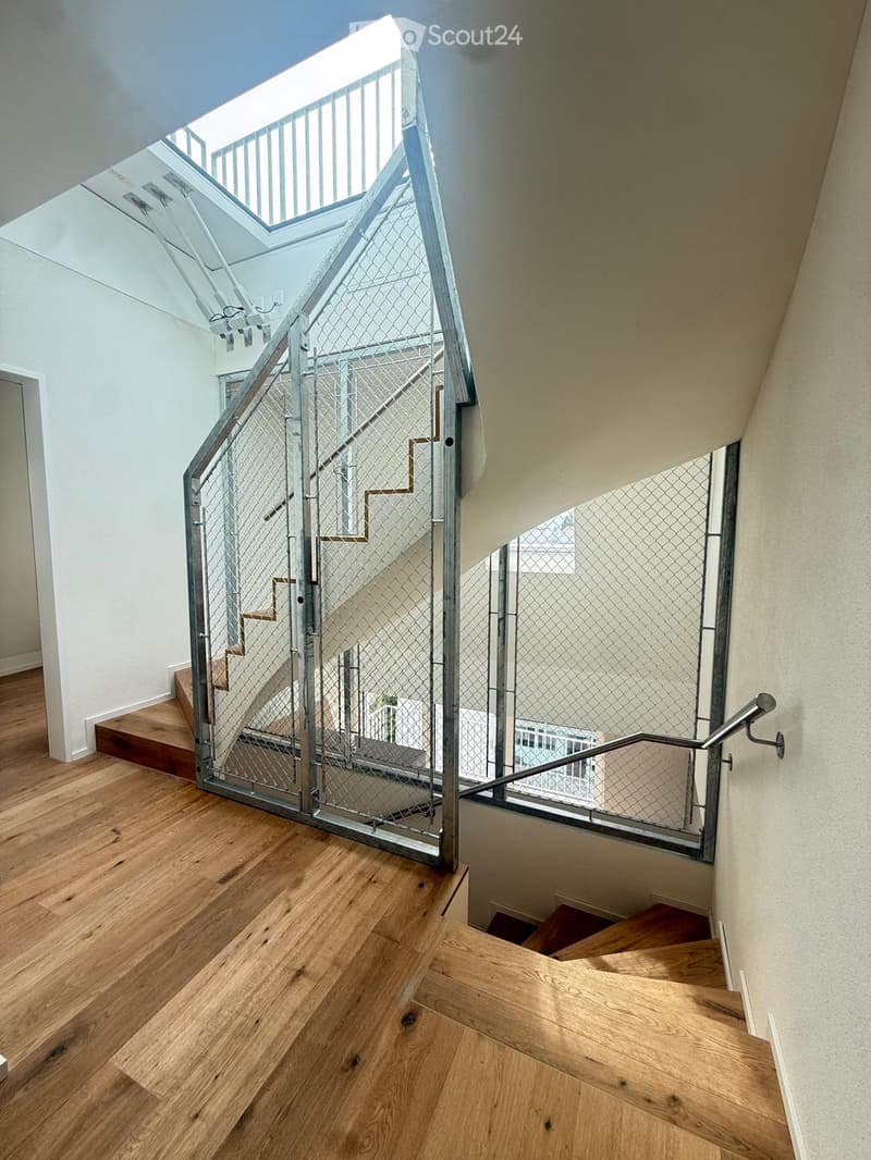 Modern staircase with wooden steps, metal railing, and glass panels, leading to an upper level with skylight windows.