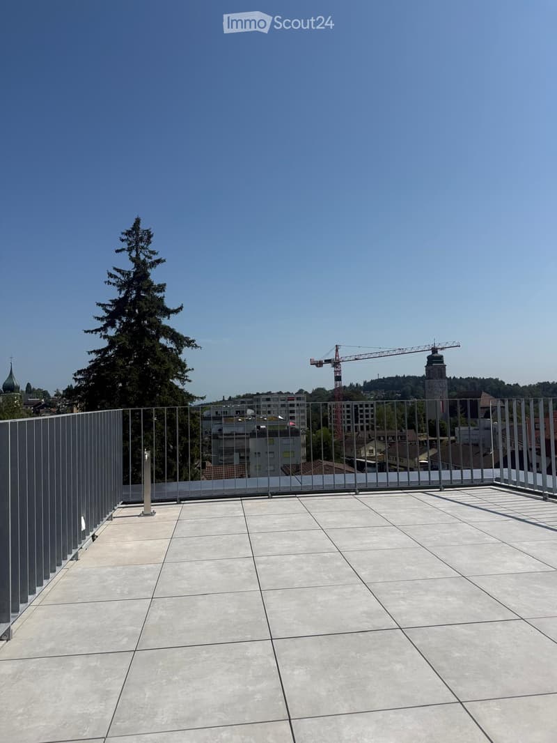 Roof terrace with modern metal railing, tiled floor, and a view of the city skyline, trees, and a construction crane.