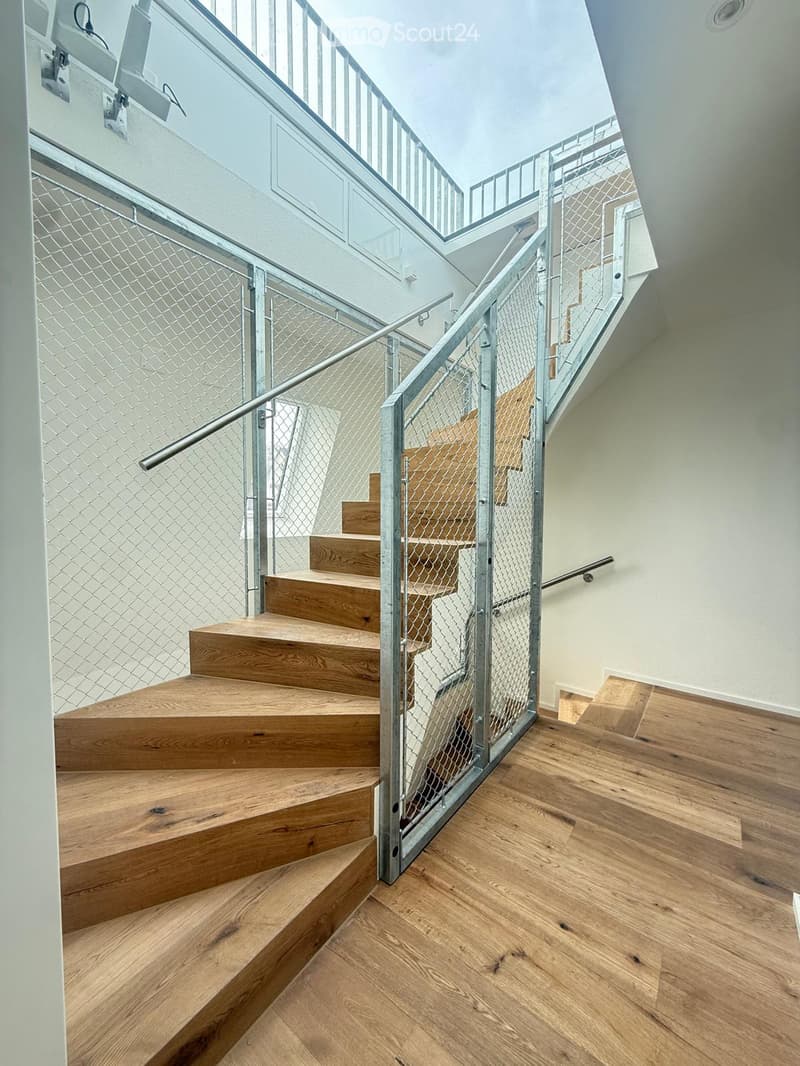 Modern staircase with wooden steps and metal railing, leading to an upper level. The hallway features light wooden flooring and white walls.