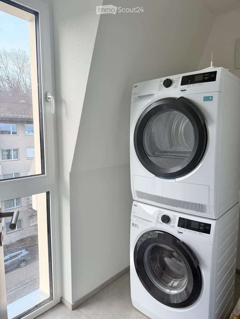 A small room with a white washing machine and dryer stacked vertically, near a window with a view of a neighboring building.