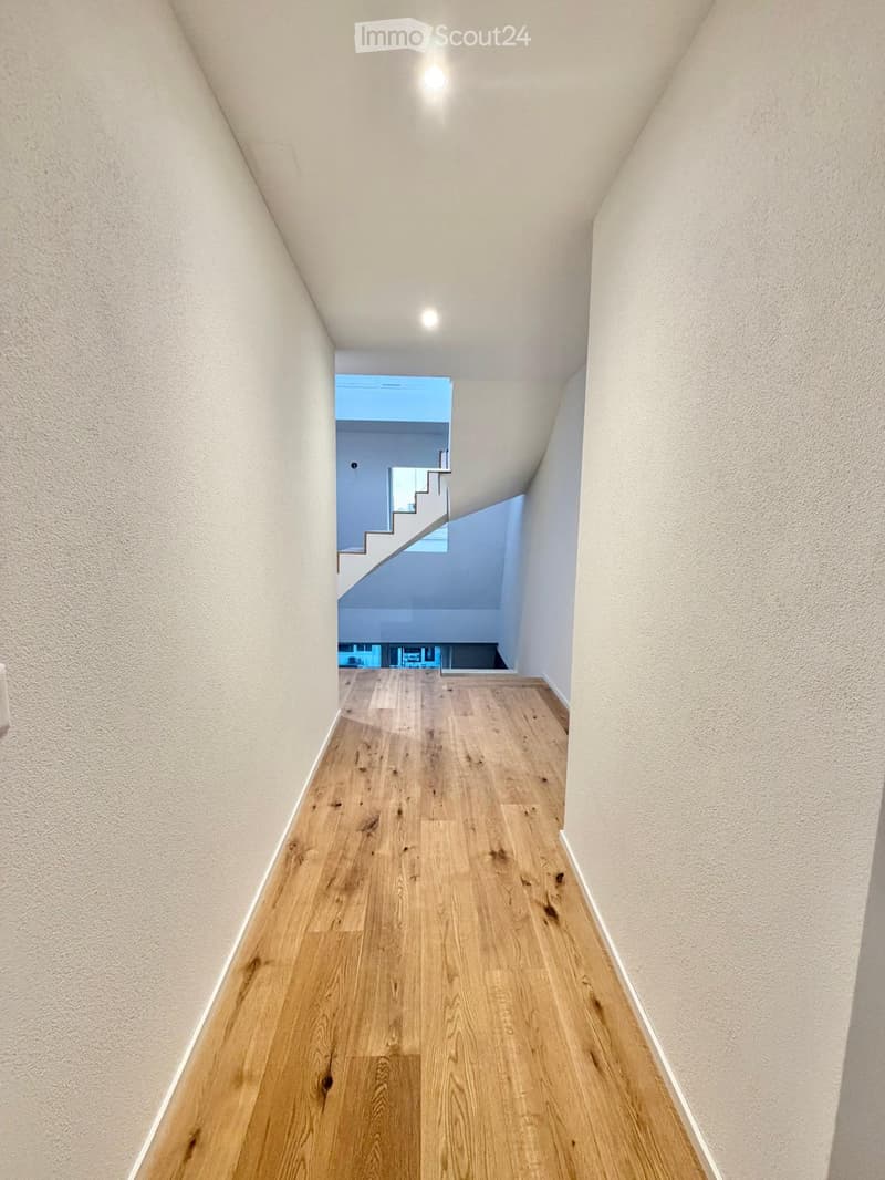 A narrow hallway with light wooden flooring and white walls, leading to a staircase area with a visible landing and railing.