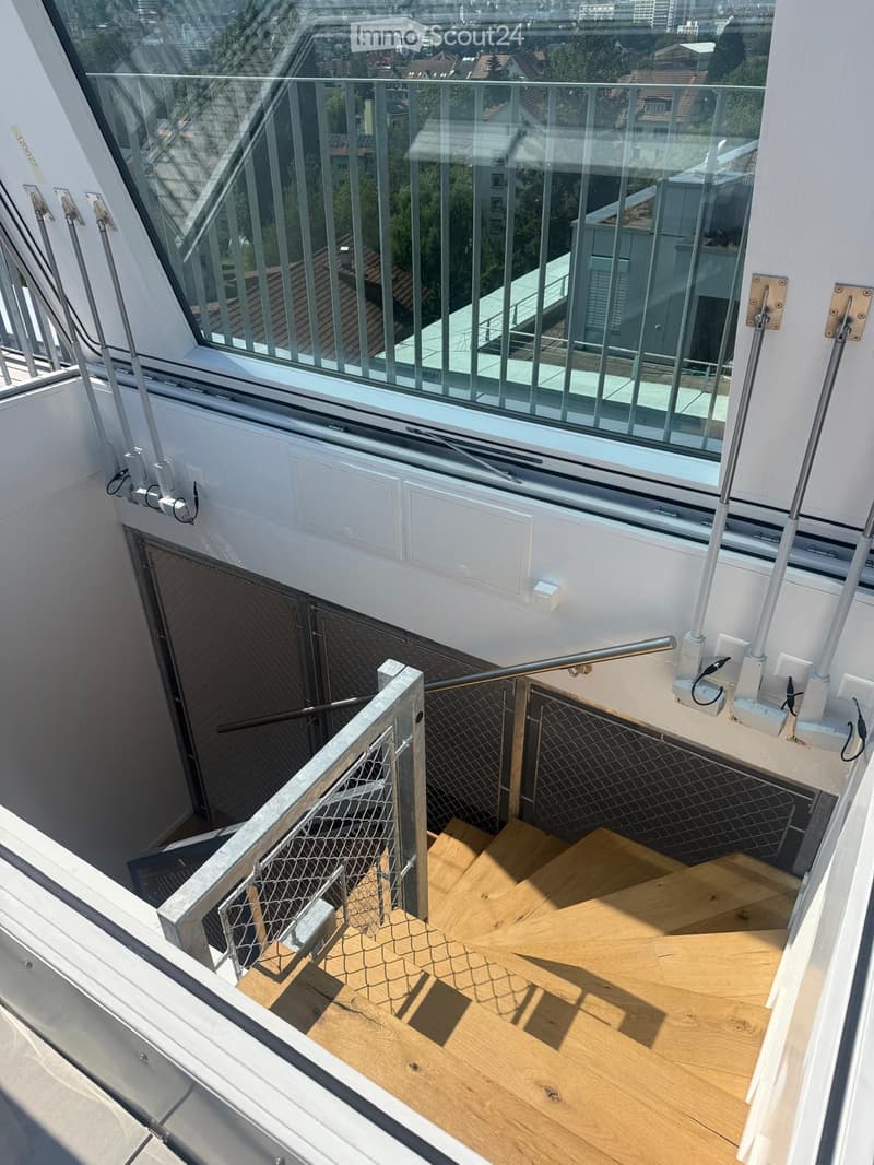 Balcony with wooden floor, metal railing, and glass panels, overlooking a cityscape with buildings and greenery.