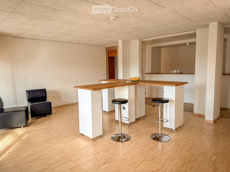 Modern, minimalist room with a wooden bar counter, two black leather sofas, and two black bar stools. The floor is light wood, and the walls are white.