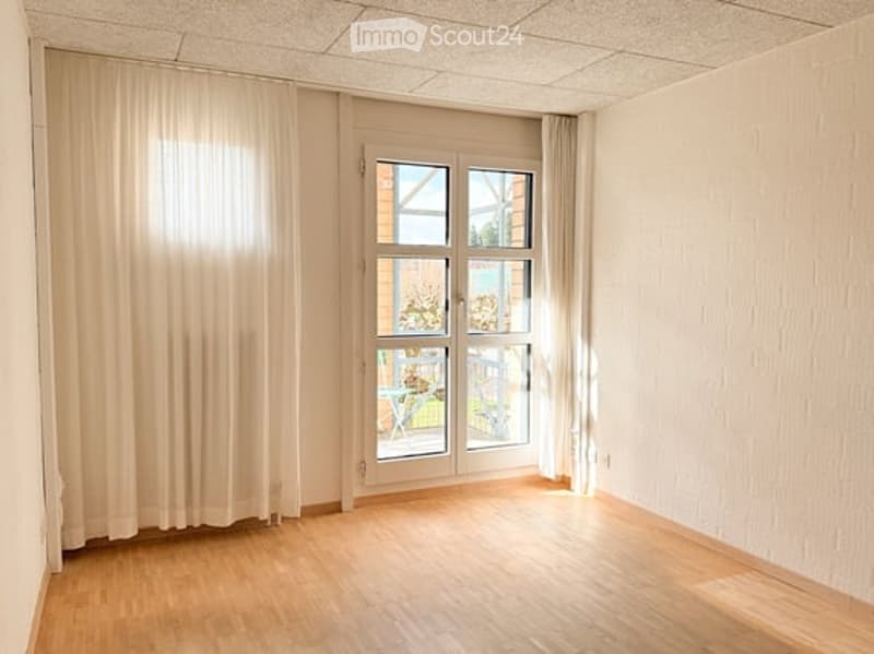 Empty room with wooden flooring, white walls, large window with white curtains, and a glass door leading to a balcony.
