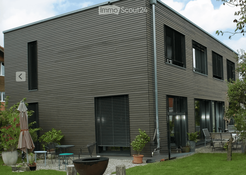 Modern house exterior with grey wooden siding, large windows, and a well-maintained garden area featuring potted plants, outdoor furniture, and a fire pit.