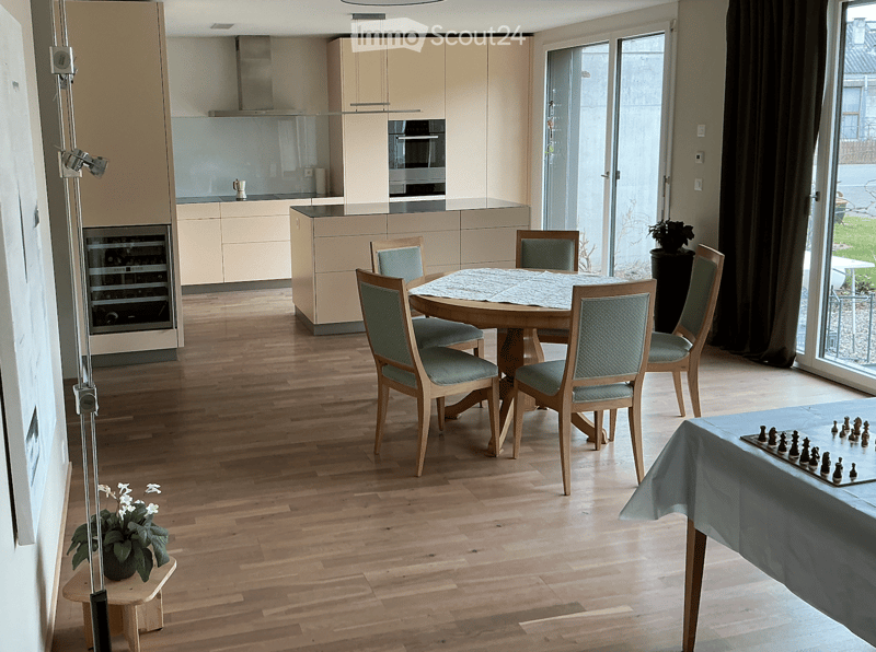 Open-plan dining and kitchen area with wooden flooring, modern light-colored cabinets, a round dining table with six chairs, a wine rack, and a chessboard on a side table.