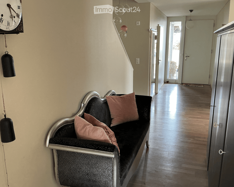 A hallway with a black sofa featuring pink pillows, a wall-mounted clock, and a wooden floor.