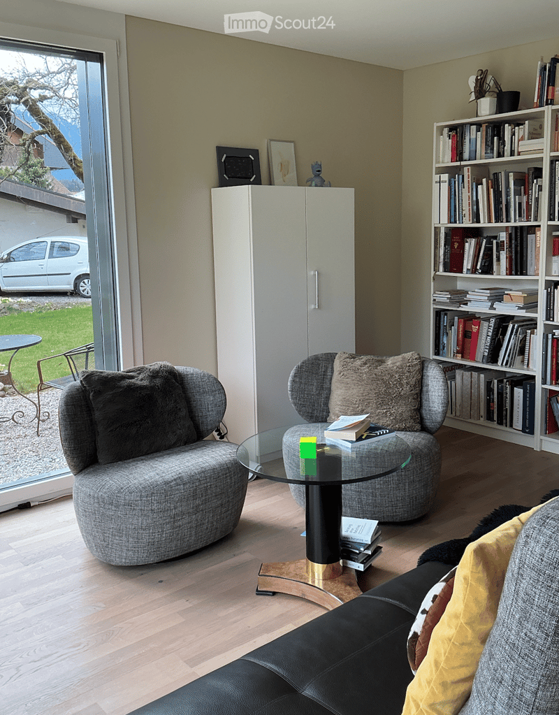 Modern living room with a large window offering a scenic view, featuring a black leather sofa, two grey armchairs, a glass coffee table, and a bookshelf filled with books.