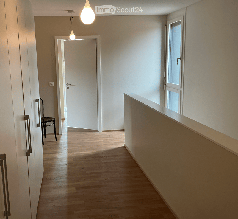 A clean, well-lit hallway with wooden flooring, white walls, and a single hanging light fixture. A black chair is positioned near the left wall, and a white door is visible at the end of the corridor.