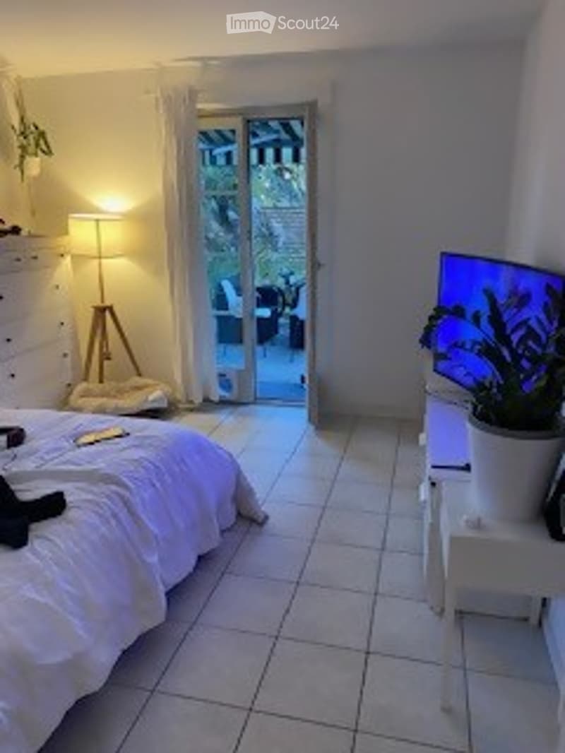 Bed, white tiled floor, bedside table with lamp, white cabinet, sliding glass door leading to balcony, potted plant, television, dog bed