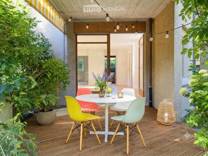 A modern outdoor patio with a white round table, colorful chairs, a vase with flowers, and string lights above.