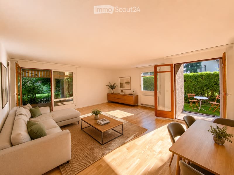 Bright living room with wooden flooring, large windows, and sliding glass doors leading to a terrace. Features a beige sectional sofa, wooden coffee table, dining table with chairs, and a wooden cabinet with decorative items.