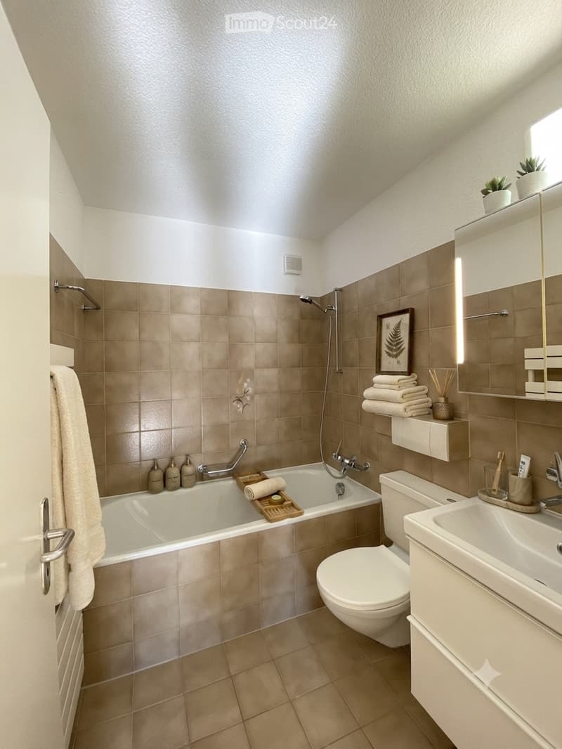Modern bathroom with beige tiles, a bathtub, toilet, sink, and a large mirror. Features include a framed picture, neatly folded towels, a wooden tray with a candle, and a small plant on the cabinet.