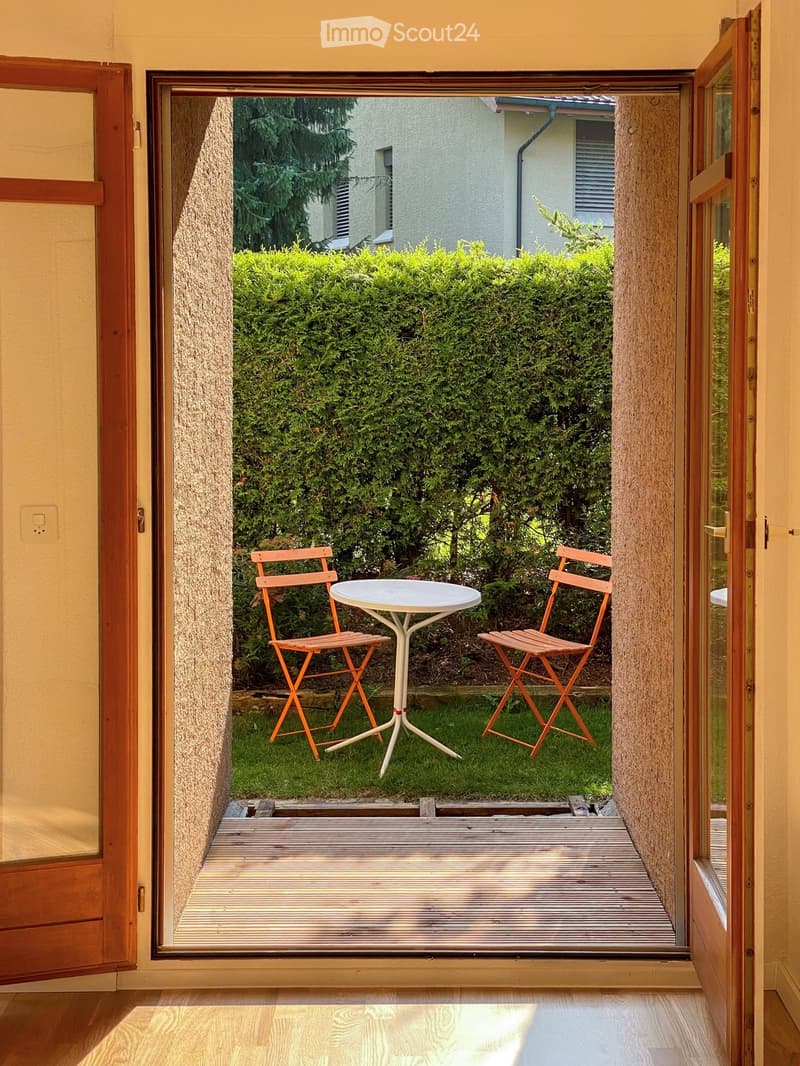 Open wooden balcony with a small table and two chairs, overlooking a green garden with bushes and trees.