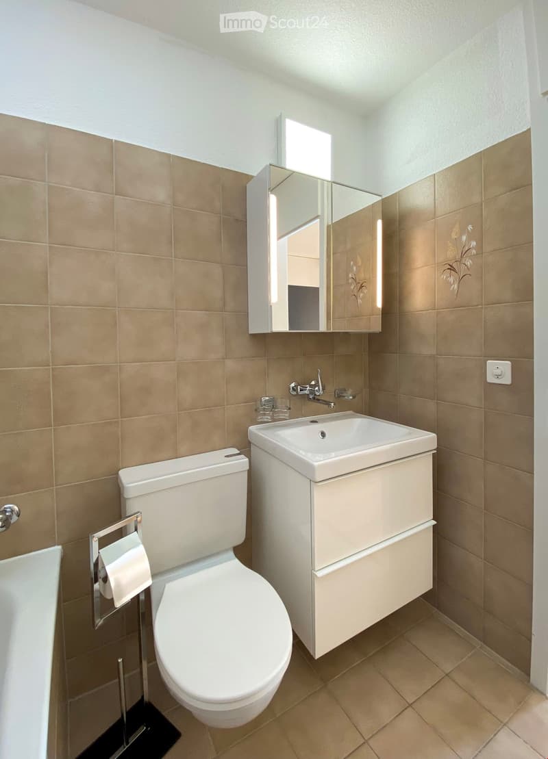 Modern bathroom with beige tiles, white toilet, sink with built-in cabinet, and illuminated mirror above the sink.