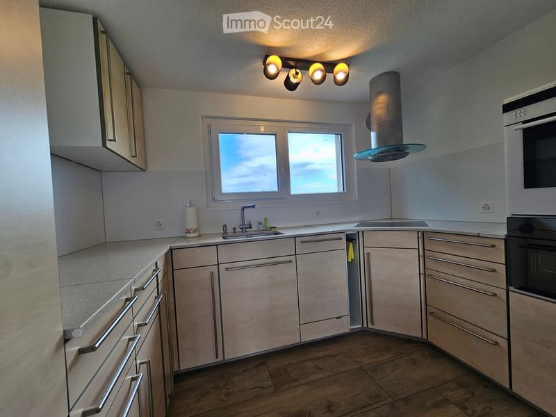 Modern kitchen with light wood cabinets, stainless steel appliances, and a central island. Features a sink, faucet, oven, and range hood with a window above.