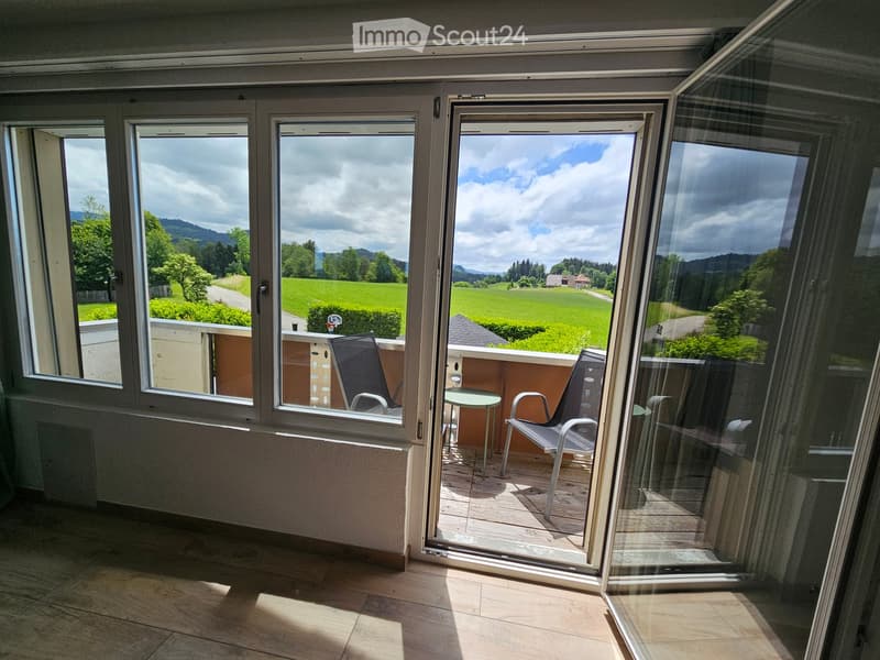 Spacious balcony with large glass doors, wooden floor, two chairs, and a small table, overlooking a green field and mountains.