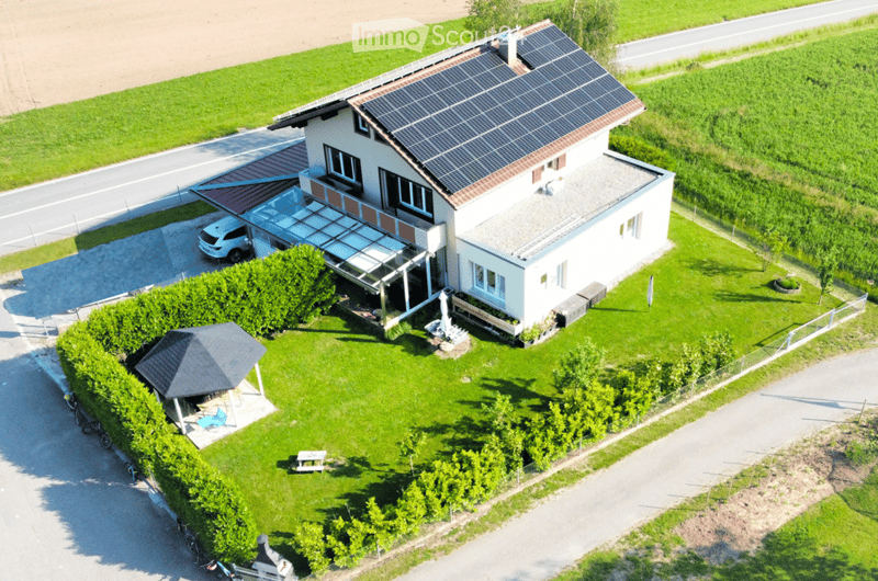 A two-story house with solar panels on the roof, surrounded by a well-maintained garden, a covered patio, and a garage. The property includes a small gazebo with outdoor furniture and a fenced yard.