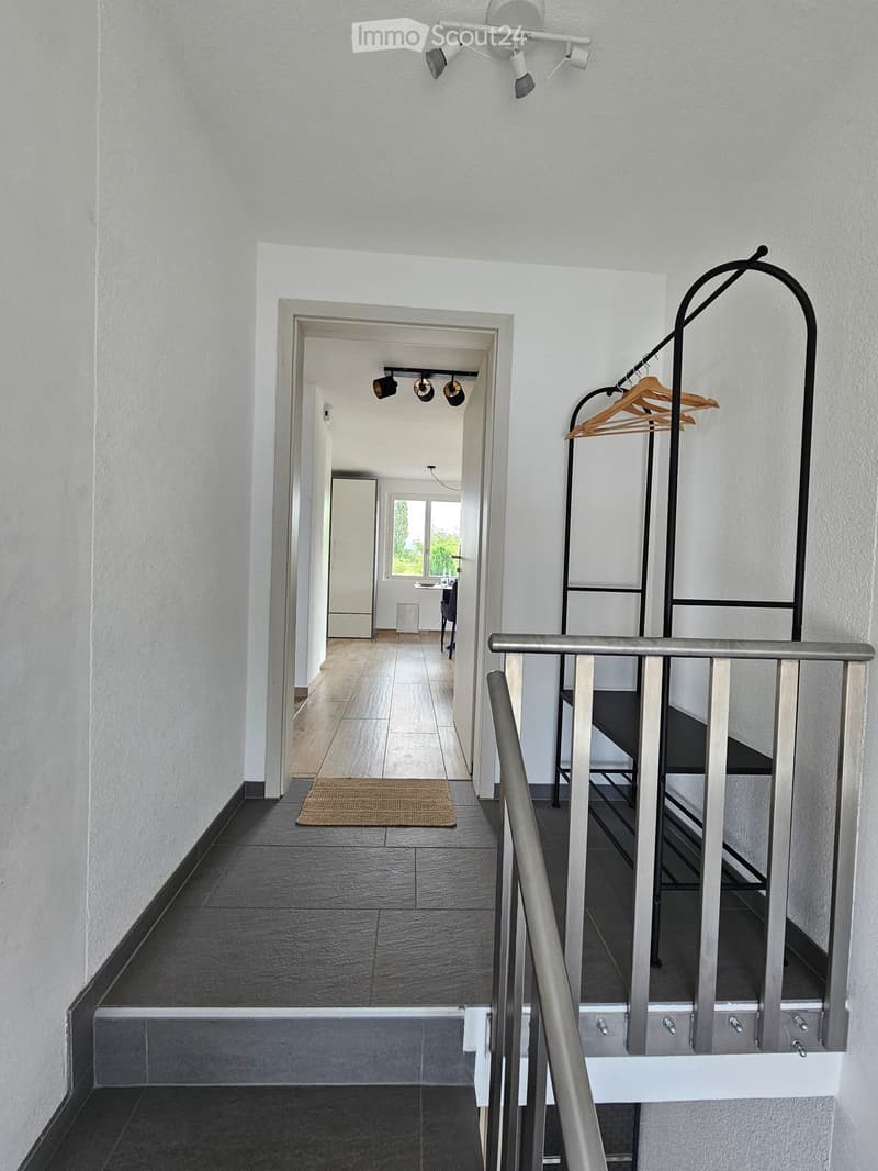 Modern hallway with grey tiled floor, white walls, and a staircase leading upwards. Features include a coat rack with hangers, a small doormat, and recessed ceiling lights.