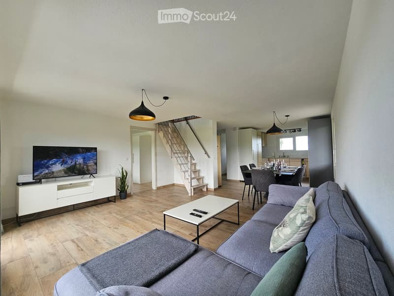 Modern living room with grey sofa, coffee table, TV, dining area, and staircase leading to another level.