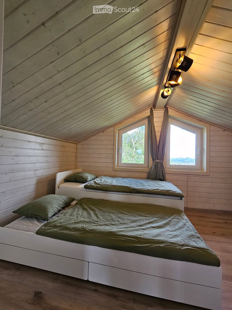 Cozy attic bedroom with wooden walls and slanted ceilings, featuring two single beds with green bedding and pillows, large windows with curtains, and warm wall-mounted lighting.