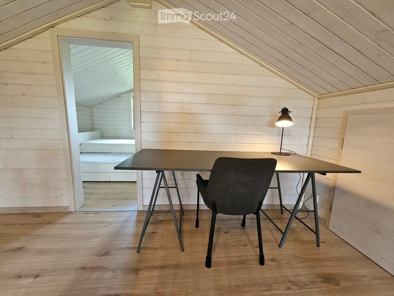 A modern, minimalist room with wooden walls and a slanted ceiling. Features a black table, a single chair, and a lamp on the table. An open doorway leads to another room with a bed visible.