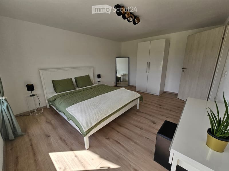 Modern bedroom with a white bed, green bedding, nightstands with lamps, a large mirror, a wardrobe, and a small table with a potted plant.