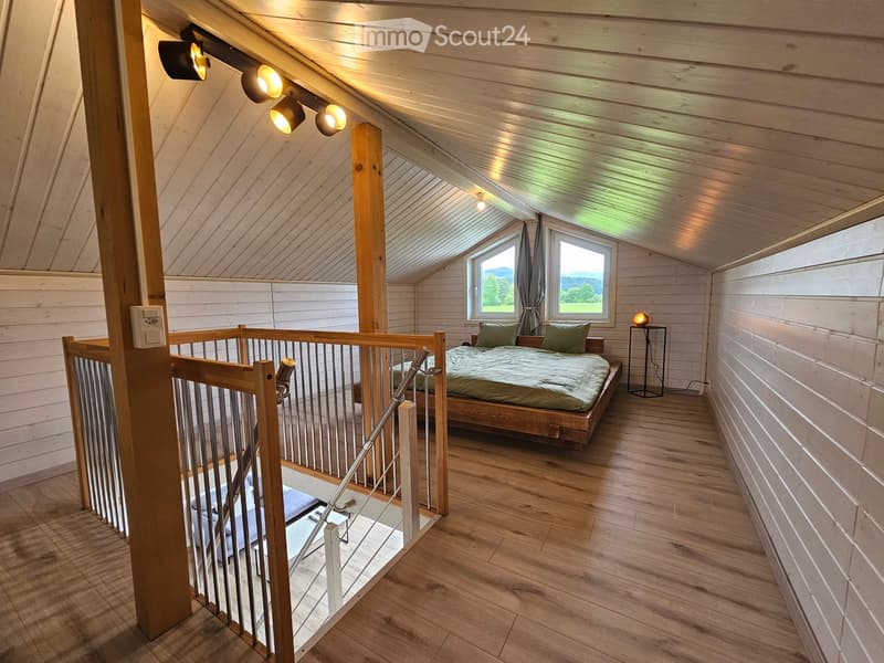 Cozy attic bedroom with wooden walls and ceiling, featuring a bed, nightstand, table lamp, and large windows with curtains.