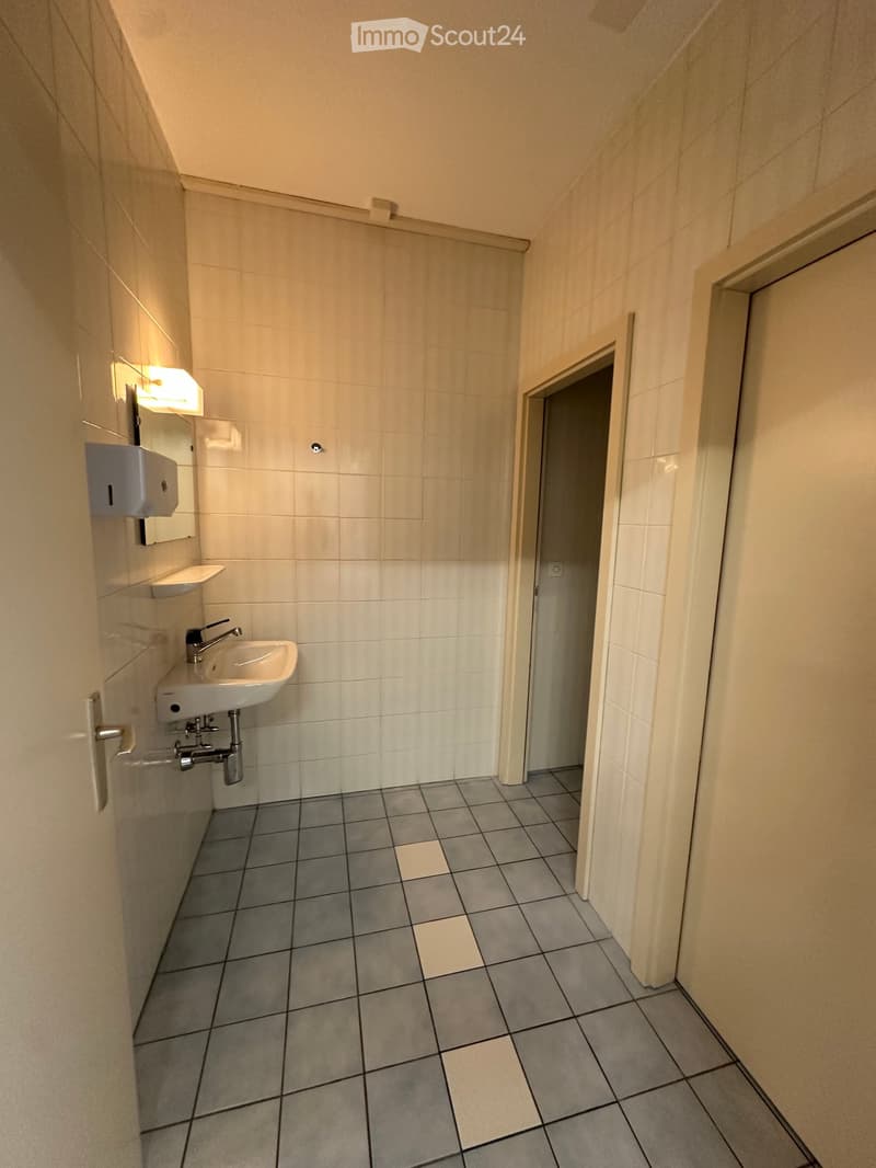 Small bathroom with a white tiled wall, a sink with a faucet, a mirror, and a light fixture above the sink. The floor is tiled in gray and white.