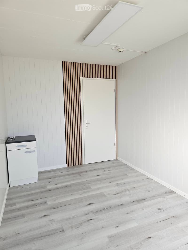 Empty room with light wooden flooring, white paneled walls, and a white cabinet with a black countertop. A white door is visible in the background.