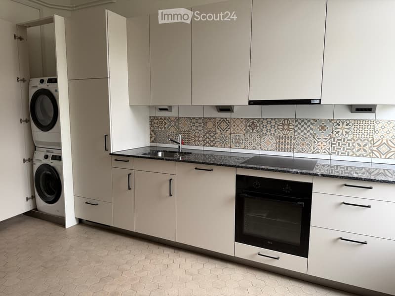 Modern kitchen with white cabinets, dark countertops, and a decorative tile backsplash. Includes a sink, faucet, oven, and a washing machine in an adjacent space.