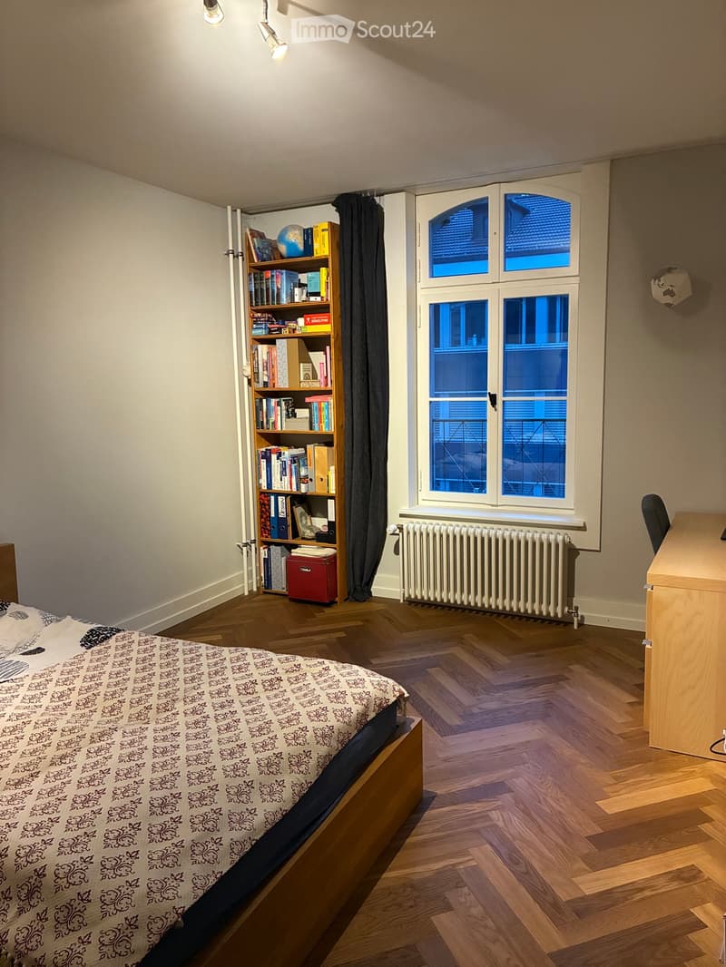 Cozy bedroom with wooden floor, large window, bed with patterned bedding, bookshelf filled with books, desk, and chair.