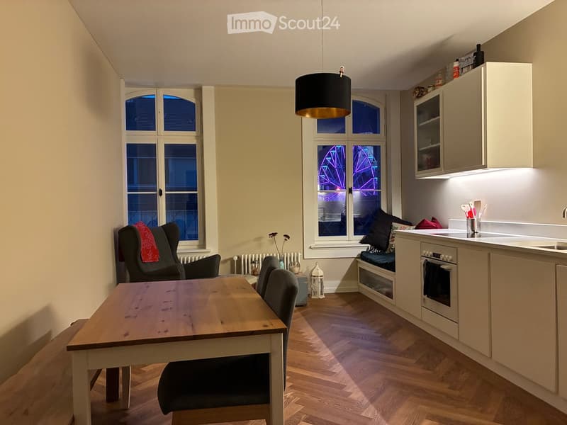 A cozy, well-lit living and dining area with a wooden table, chairs, a sofa, and a small kitchen area featuring cabinets, an oven, and a sink.