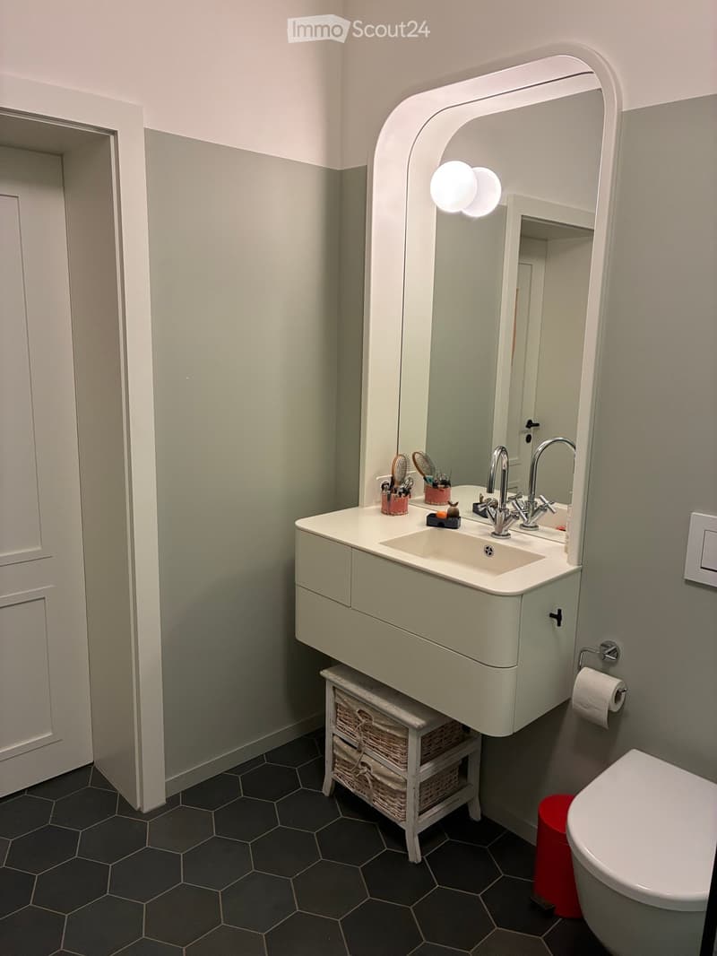 Modern bathroom with a white sink, chrome faucet, large arched mirror with built-in lighting, wicker basket storage, and a toilet. The walls are painted in a two-tone grey scheme, and the floor features hexagonal tiles.