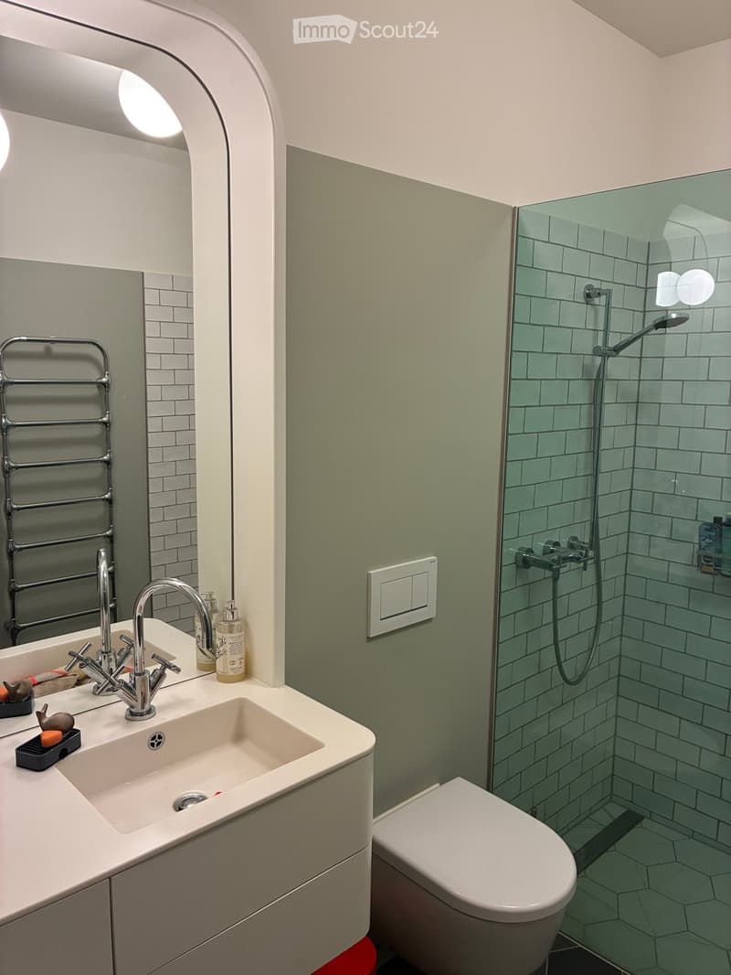 Modern bathroom with a white sink, chrome faucet, large mirror, toilet, and a glass-enclosed shower with green tiles.