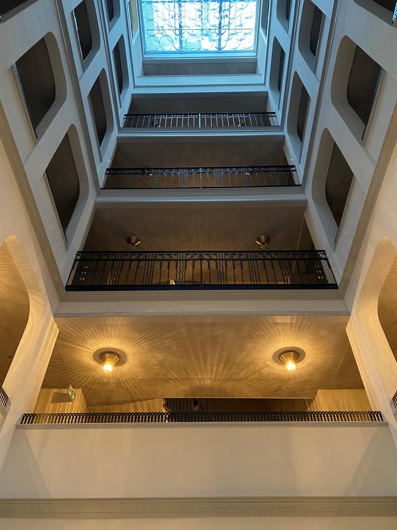 Elegant interior view of a multi-level atrium with ornate metal railings, warm lighting, and a skylight at the top.