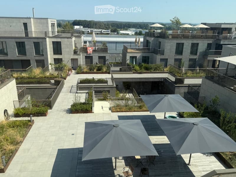 Rooftop garden area with tiled walkways, wooden decking, several large umbrellas, chairs, and tables, surrounded by low greenery and modern buildings.
