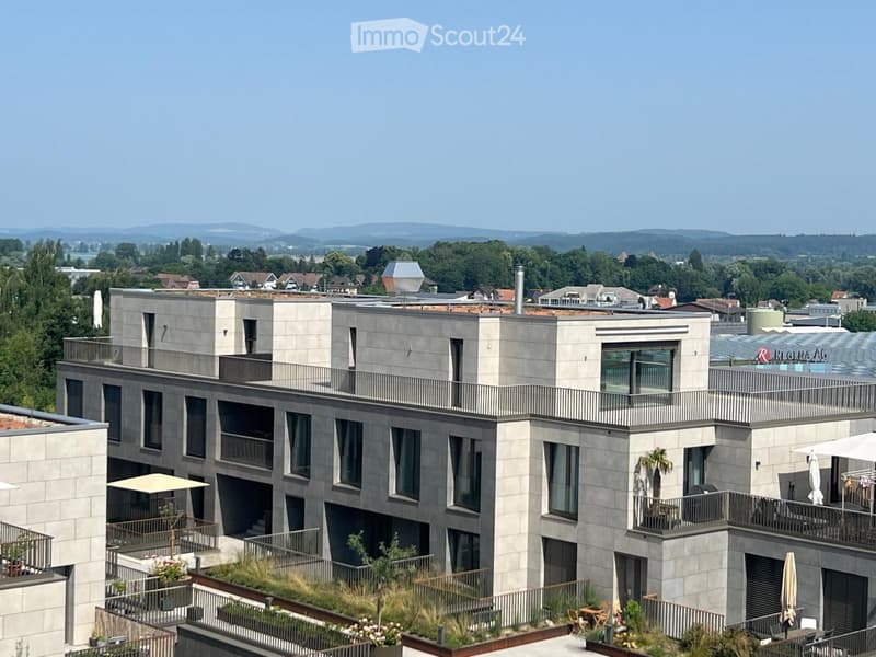 Modern multi-story building with grey stone facade, large windows, balconies, and rooftop garden area with outdoor furniture and greenery.
